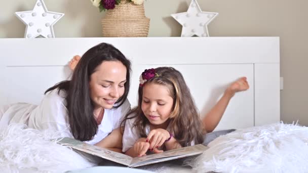 Mamá lee un libro con su hija. Niña, la colegiala está leyendo en el dormitorio de la cama. Comunicación entre hijos y padres. Colores diurnos y claros en el marco. Enfoque suave. — Vídeo de stock