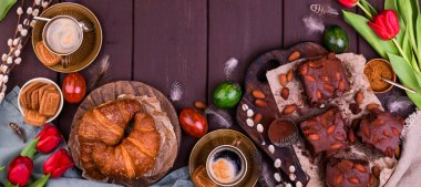 Paskalya kahvaltısında aromatik taze espresso, kruvasan, kek, renkli yumurta, kırmızı lale ve söğüt. Kurabiyeli kahve, ahşap masadaki çiçekler. Yukarıdan görüntüle.