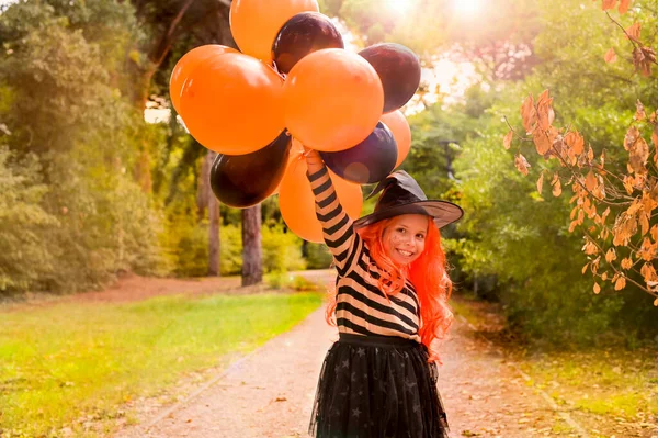 Happy holiday Halloween. Little cheerful girl in a witch costume and colored balloons. Sun glare in the frame. Soft focus.
