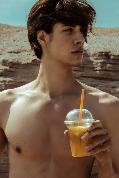 Strong face commercial wet male model with lemonade in swimwear on the cloudy sky and sand background