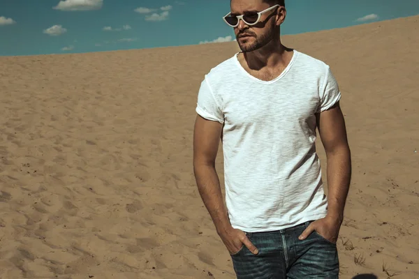 Strong commercial face male model  wearing white sunglesses jeans and white t-shirt  on sand and  blue sky with clouds