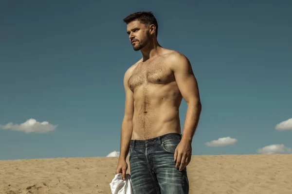 Strong commercial face male model  wearing white sunglesses jeans and white t-shirt  on sand and  blue sky with clouds