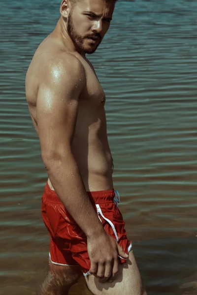 夏の感じと水の背景に赤い水着ショート パンツでセクシーな強いひげを生やした男性塊 — ストック写真
