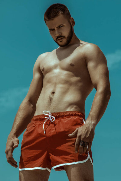 Sexy strong bearded male hunk in red swim shorts on the water background with summer vibes