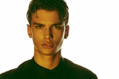 Siyah gömlekli, siyah pantolonlu, beyaz izole font arka planda sarı ışık altında güçlü kaslı erkek modeli.