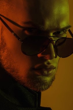 Kas gücü yüzlü, güzel, soyulmuş, sıradan görünümlü, kot pantolon ve takım elbiseli, yeşil gözlüklü, bej renkli, izole font arka planda sarı ışık takan erkek model.