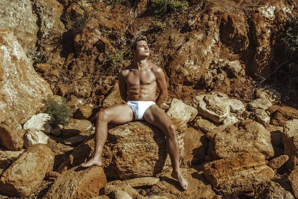 Strong face male model in white underwear on the nature background with sand, rocks and water on the beach on the sunset