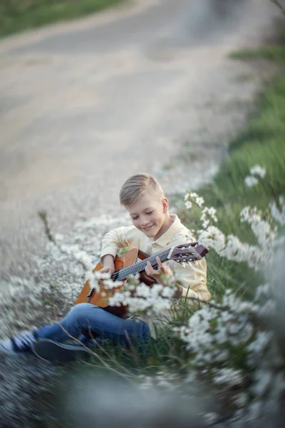 Außenporträt eines jungen Teenagers, der Gitarre spielt und in der Nähe eines Parks auf der Straße sitzt. — Stockfoto