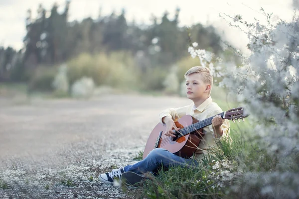 Outdoor Portret van een jonge tiener gitaarspelen en zittend op de weg in de buurt van park. — Stockfoto