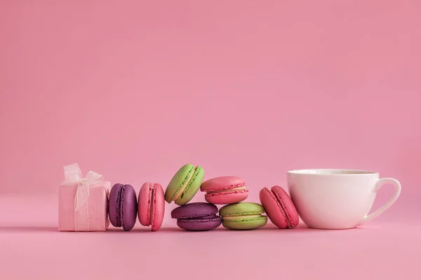 Pink present and Row of french colorful macaroons and cup of coffee, lying on pink background