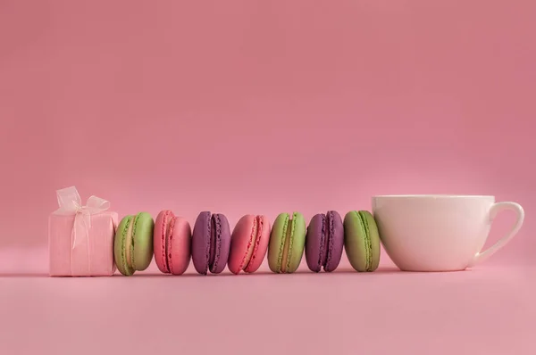 Pink present and Row of french colorful macaroons and cup of coffee, lying on pink background