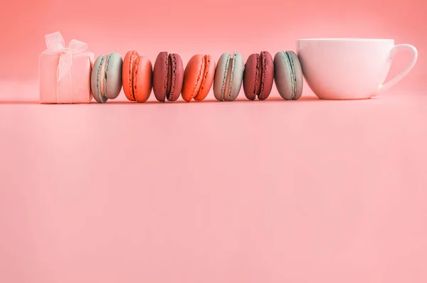 Розовый подарок и ряд французских красочных макарон и чашку кофе, лежащих на коралловом фоне — стоковое фото