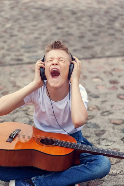 Adolescente escuchando música en los auriculares y gritando canción. Joven sentado con una guitarra en la calle . —  Fotos de Stock