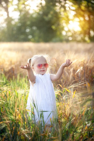 Küçük kız güneşli yaz alanında oynuyor. Beyaz elbise ve güneş gözlüğü bebek. — Stok fotoğraf