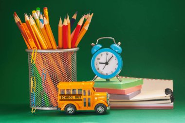 Eğitim ve okul kavramına geri dönelim. Sarı retro okul otobüsü ve sepet, ders kitapları ve koyu yeşil arka plan üzerinde çalar saat kalemler.