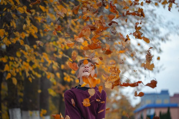 Przystojny szczęśliwy chłopiec rzuca opadłe liście w górę, bawiąc się w jesiennym parku. — Zdjęcie stockowe