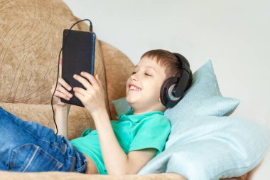 Çocuk oturma odasındaki koltukta dijital tablet ve kulaklık kullanıyor. Çocuk evden ders çalışıyor ve aletlerle oynuyor.