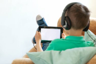 Çocuk dijital tablet ve kulaklık kullanıyor. Oturma odasında beyaz ekran kanepesi var..