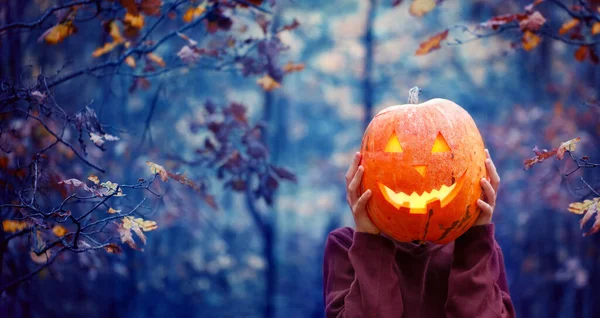 Fiú kezében faragott tök halloween előtt a fejét a sötét őszi erdőben. Halloween ünnep koncepció. — Stock Fotó