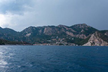 Sakin mavi deniz ve gökyüzü, arkasında dağlar ve küçük bir kasaba var.
