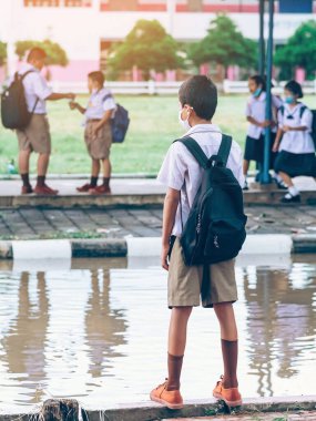 Erkek ilkokul öğrencisi Coronavirus 'u (Covid-19) önlemek için yüz maskesi takıyor ailesinin okuldan sonra eve dönmesini bekliyor ve okul kapısının önünde yağmur duruyor.