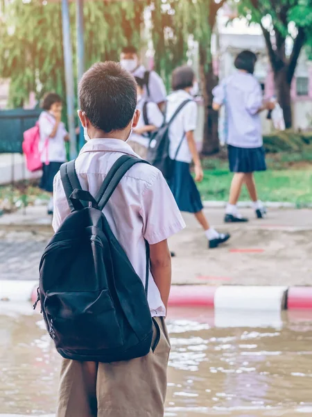 Male elementary school student wear face mask to prevent the Coronavirus(Covid-19) wait for her parents to pick her up to return home after school and the rain just stop in front of the school gate