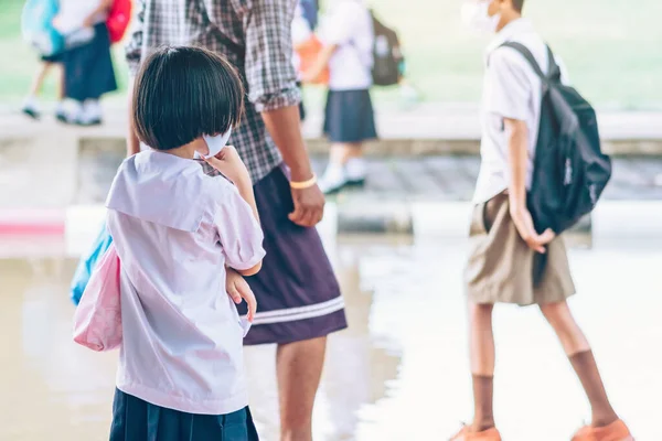 Female elementary school student wear face mask to prevent the Coronavirus(Covid-19) wait for her parents to pick her up to return home after school and the rain just stop in front of the school gate