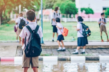 Erkek ilkokul öğrencisi Coronavirus 'u (Covid-19) önlemek için yüz maskesi takıyor ailesinin okuldan sonra eve dönmesini bekliyor ve okul kapısının önünde yağmur duruyor.