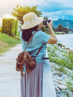 Corona Virüsü 'nü (Covid-19) öldürmek için mini taşınabilir alkol jeli şişesi günbatımından sonra nehrin yanında fotoğraf çekmek için akıllı telefon kullanan bir kadının deri omuz çantasına asılır. Yeni normal yaşam tarzı.