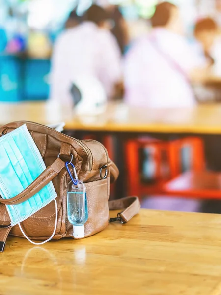 Mini portable alcohol gel bottle to kill Corona Virus(Covid-19) hang on a leather shoulder bag of a woman with a surgical mask on table at cafeteria.New normal lifestyle.Selective focus on alcohol gel