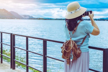 Corona Virüsü 'nü (Covid-19) öldürmek için mini taşınabilir alkol jeli şişesi günbatımından sonra nehrin yanında fotoğraf çekmek için akıllı telefon kullanan bir kadının deri omuz çantasına asılır. Yeni normal yaşam tarzı.