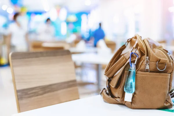 Mini portable alcohol gel bottle to kill Corona Virus(Covid-19) hang on a leather shoulder bag of a woman on table at cafeteria. New normal lifestyle.Selective focus on alcohol gel