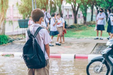 Erkek ilkokul öğrencisi Coronavirus 'u (Covid-19) önlemek için yüz maskesi takıyor ailesinin okuldan sonra eve dönmesini bekliyor ve okul kapısının önünde yağmur duruyor.