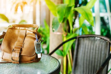 Corona Virüsü 'nü (Covid-19) öldürmek için taşınabilir mini alkol jeli şişesi bahçedeki masada kahverengi deri bir omuz çantasına asılır. Yeni normal yaşam tarzı. Sağlık hizmeti konsepti. Alkol jeline odaklan