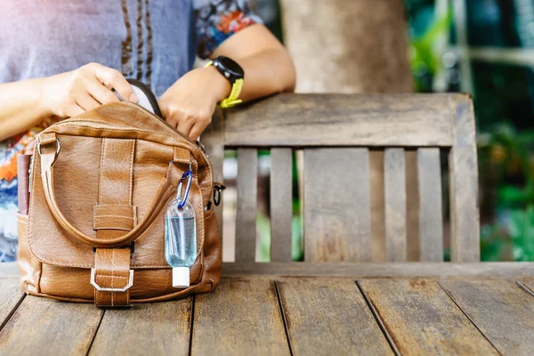 Mini portable alcohol gel bottle to kill Corona Virus(Covid-19) hang on a brown leather shoulder bag on table in garden. New normal lifestyle. Health care concept. Selective focus on alcohol gel