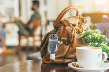 Corona Virüsü 'nü (Covid-19) öldürmek için mini alkol jeli şişesi kafede üzerinde sıcak kahve olan kahverengi deri bir omuz çantasına asılır. Yeni normal yaşam tarzı. Sağlık hizmetleri kavramı. Alkol jeline odaklanın.