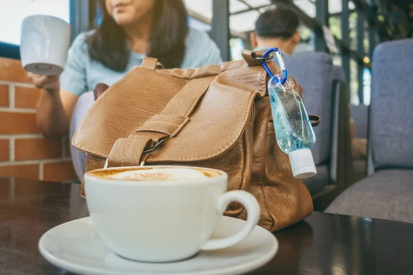 Mini alcohol gel bottle to kill Corona Virus(Covid-19) hang on a brown leather shoulder bag with hot coffee on table in cafe.New normal lifestyle.Health care concept.Selective focus on alcohol gel