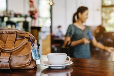 Corona Virüsü 'nü (Covid-19) öldürmek için mini alkol jeli şişesi kafede üzerinde sıcak kahve olan kahverengi deri bir omuz çantasına asılır. Yeni normal yaşam tarzı. Sağlık hizmetleri kavramı. Alkol jeline odaklanın.