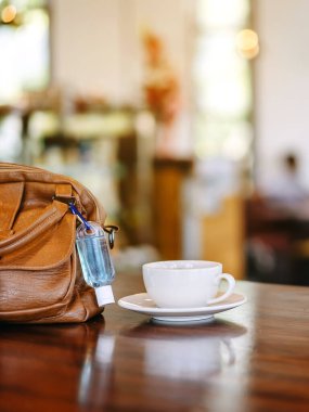 Corona Virüsü 'nü (Covid-19) öldürmek için mini alkol jeli şişesi kafede üzerinde sıcak kahve olan kahverengi deri bir omuz çantasına asılır. Yeni normal yaşam tarzı. Sağlık hizmetleri kavramı. Alkol jeline odaklanın.