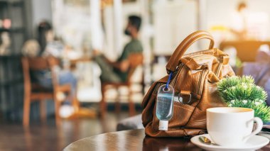 Corona Virüsü 'nü (Covid-19) öldürmek için mini alkol jeli şişesi kafede üzerinde sıcak kahve olan kahverengi deri bir omuz çantasına asılır. Yeni normal yaşam tarzı. Sağlık hizmetleri kavramı. Alkol jeline odaklanın.