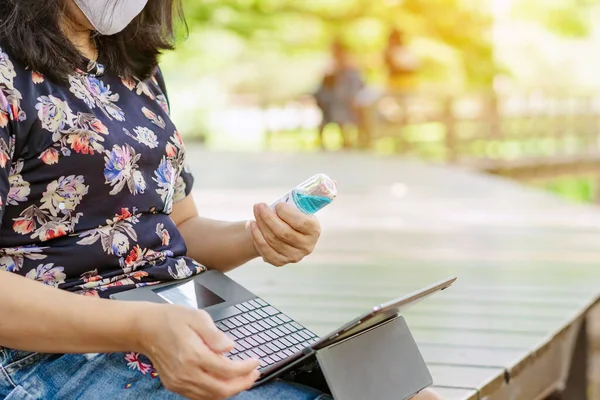 Woman wears protective mask using alcohol nano mist sprayer cleaning her hands to prevent the Coronavirus(Covid-19) and bacterias before use laptop in garden.New normal lifestyle.Health care concept.
