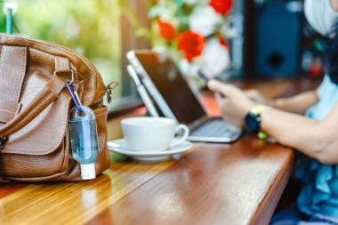 Corona Virüsü 'nü (Covid-19) öldürmek için mini alkol jeli şişesi kafede üzerinde sıcak kahve olan kahverengi deri bir omuz çantasına asılır. Yeni normal yaşam tarzı. Sağlık hizmetleri kavramı. Alkol jeline odaklanın.