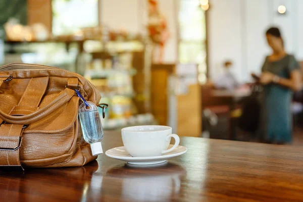 Mini alcohol gel bottle to kill Corona Virus(Covid-19) hang on a brown leather shoulder bag with hot coffee on table in cafe.New normal lifestyle.Health care concept.Selective focus on alcohol gel