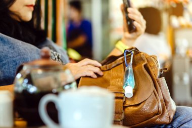 Corona Virüsü 'nü (Covid-19) öldürmek için mini taşınabilir alkol jeli şişesi kafede sıcak kahveli kahverengi deri bir omuz çantasına asılır. Yeni normal yaşam tarzı. Sağlık hizmeti kavramı. Alkol jeline odaklı.