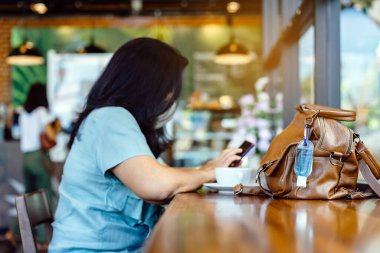 Corona Virüsü 'nü (Covid-19) öldürmek için mini alkol jeli şişesi kafede üzerinde sıcak kahve olan kahverengi deri bir omuz çantasına asılır. Yeni normal yaşam tarzı. Sağlık hizmetleri kavramı. Alkol jeline odaklanın.