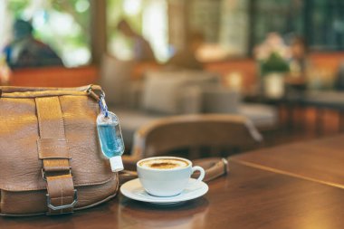 Corona Virüsü 'nü (Covid-19) öldürmek için mini alkol jeli şişesi kafede üzerinde sıcak kahve olan kahverengi deri bir omuz çantasına asılır. Yeni normal yaşam tarzı. Sağlık hizmetleri kavramı. Alkol jeline odaklanın.