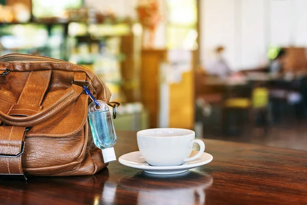 Corona Virüsü 'nü (Covid-19) öldürmek için mini alkol jeli şişesi kafede üzerinde sıcak kahve olan kahverengi deri bir omuz çantasına asılır. Yeni normal yaşam tarzı. Sağlık hizmetleri kavramı. Alkol jeline odaklanın.