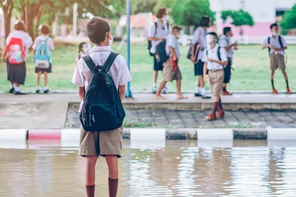 Male elementary school student wear face mask to prevent the Coronavirus(Covid-19) wait for her parents to pick her up to return home after school and the rain just stop in front of the school gate