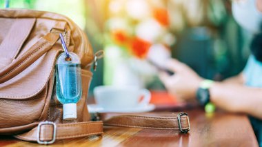 Corona Virüsü 'nü (Covid-19) öldürmek için mini alkol jeli şişesi kafede üzerinde sıcak kahve olan kahverengi deri bir omuz çantasına asılır. Yeni normal yaşam tarzı. Sağlık hizmetleri kavramı. Alkol jeline odaklanın.