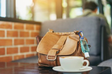 Corona Virüsü 'nü (Covid-19) öldürmek için mini alkol jeli şişesi kafede üzerinde sıcak kahve olan kahverengi deri bir omuz çantasına asılır. Yeni normal yaşam tarzı. Sağlık hizmetleri kavramı. Alkol jeline odaklanın.